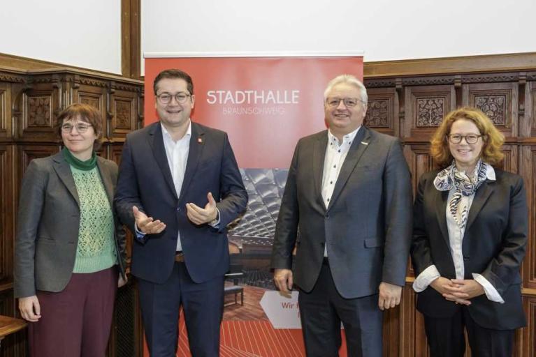 Vier Personen stehen in einem holzvertäfelten Raum vor einem Banner der Stadthalle Braunschweig