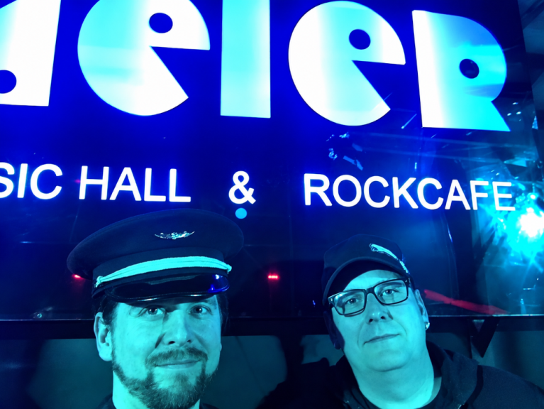 Zwei Männer stehen vor einer leuchtenden Club- oder Konzertbeschriftung mit den Worten Music Hall & Rockcafe.