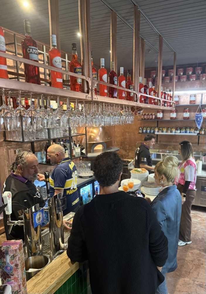 Menschen stehen in einer Bar an der Theke, darüber hängen Gläser und Flaschen mit roten Spirituosen.