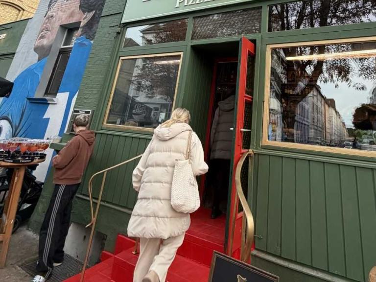 Eingang einer Pizzeria mit grünem Fassadenschild, roter Treppe und Personen vor der Tür