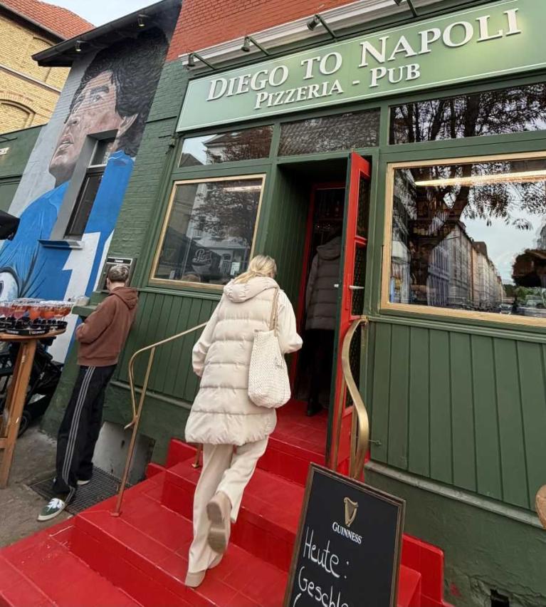 Eingang einer Pizzeria mit grünem Fassadenschild, roter Treppe und Personen vor der Tür