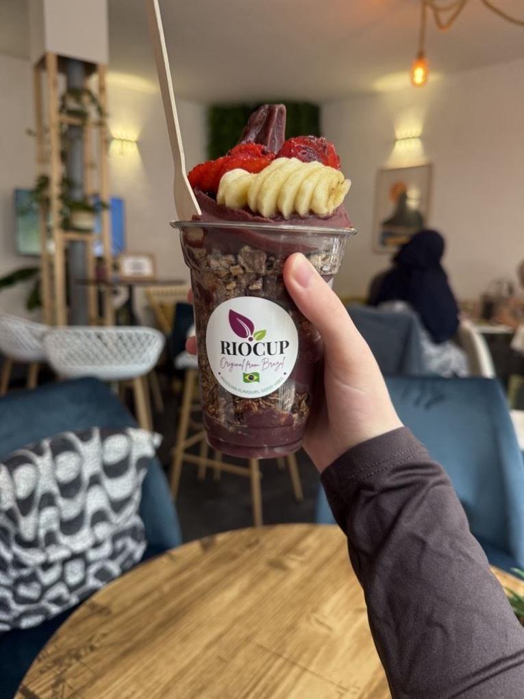 Eine Hand hält einen Becher mit Acai-Bowl und Früchten in einem Café