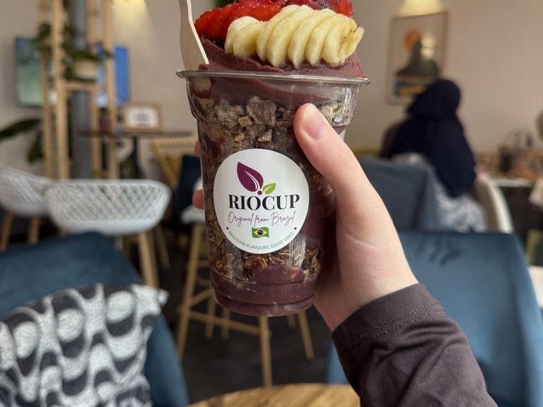 Eine Hand hält einen Becher mit Acai-Bowl und Früchten in einem Café