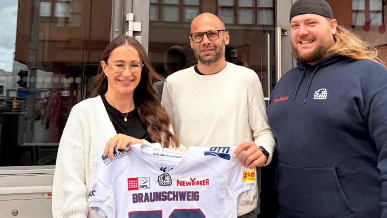 Drei Personen stehen vor einem Gebäude und halten ein Braunschweig-Trikot