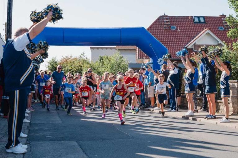 Kinder und Erwachsene laufen bei einem Straßenlauf unter einem blauen Zielbogen, flankiert von Cheerleadern mit Pompons.
