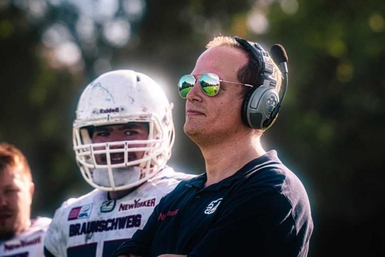 Ein Mann mit Sonnenbrille und Headset steht vor einem Footballspieler mit Helm.