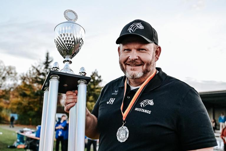 Ein Mann in schwarzer Mustang-Kleidung hält einen großen Pokal im Freien.