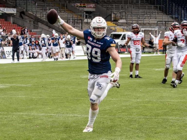 Footballspieler in blauem Trikot jubelt mit erhobener Hand und Ball auf dem Spielfeld