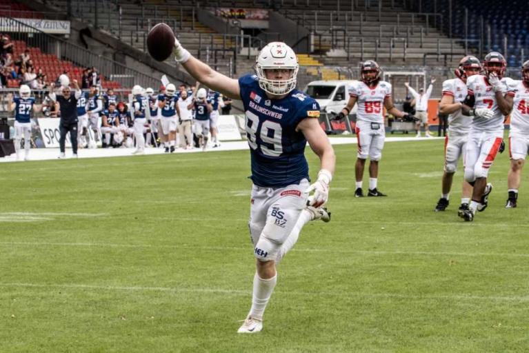 Footballspieler in blauem Trikot jubelt mit erhobener Hand und Ball auf dem Spielfeld