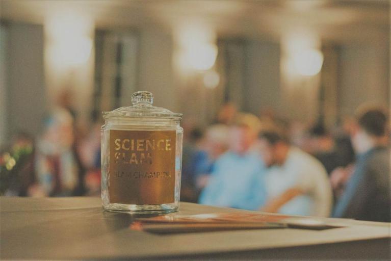 Ein Glasbehälter mit der Aufschrift Science Jam steht auf einem Tisch, im Hintergrund sitzen unscharf mehrere Personen.