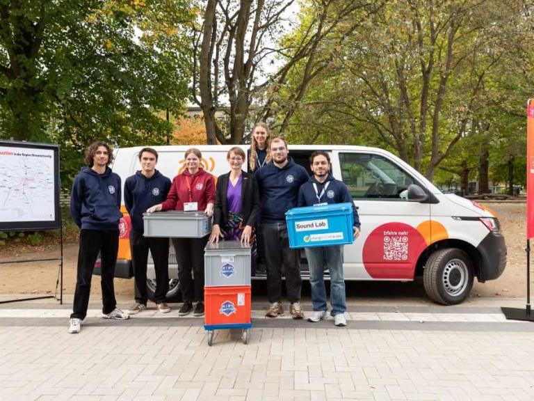 Eine Gruppe von sechs Personen steht vor einem weißen Transporter und hält mehrere Kisten im Freien.