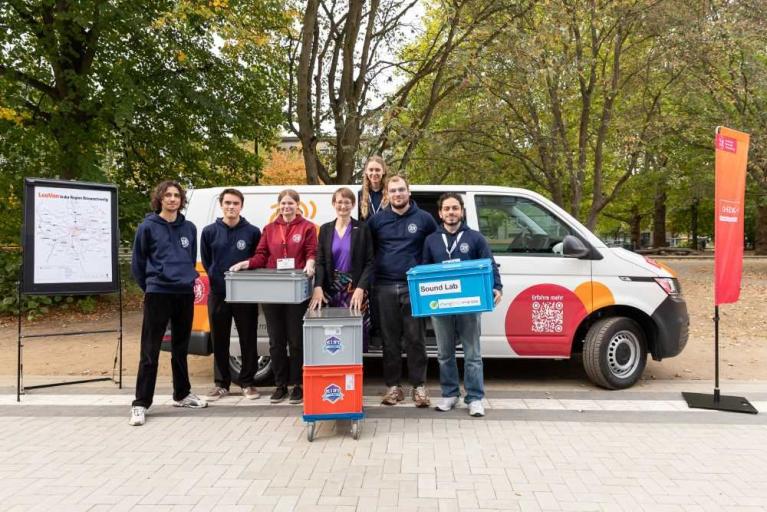 Eine Gruppe von sechs Personen steht vor einem weißen Transporter und hält mehrere Kisten im Freien.