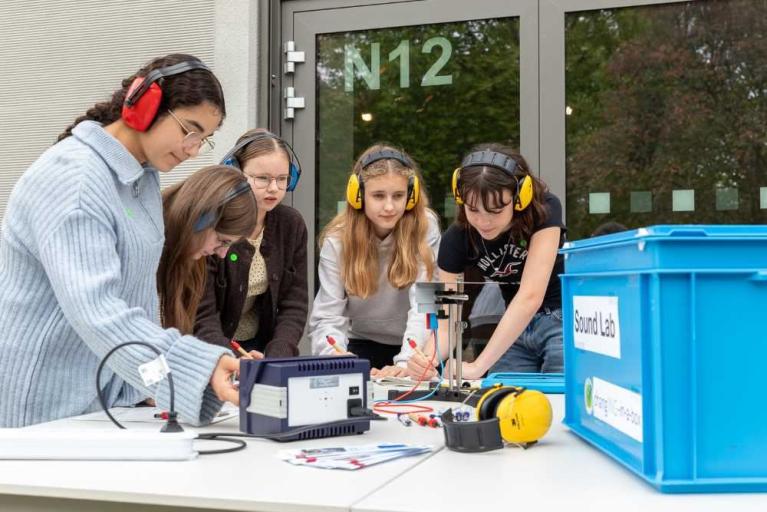 Eine Gruppe von Kindern und eine erwachsene Frau arbeitet mit Elektronik und Messgeräten an einem Tisch, alle tragen Kopfhörer.