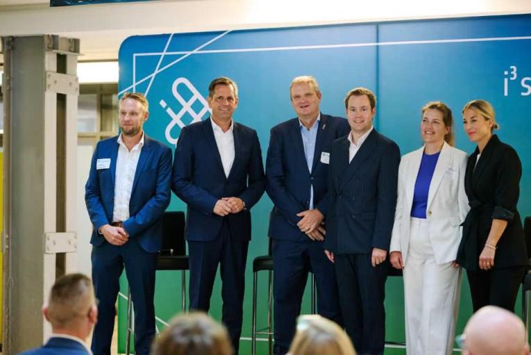 Sechs Personen in Anzügen und Businesskleidung stehen auf einer Bühne vor einem blauen Hintergrund.