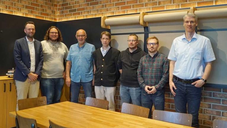 Sieben Männer stehen gemeinsam in einem Raum vor einer Backsteinwand und einer Projektionswand.