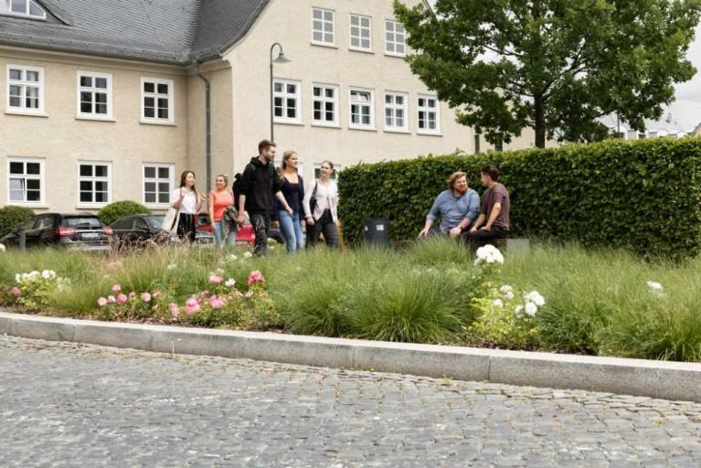 Mehrere junge Erwachsene gehen und sitzen in einem begrünten Innenhof vor einem Gebäude.