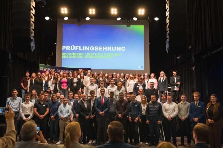 Große Gruppe von Menschen auf einer Bühne bei einer Prüfungsfeier vor einer Leinwand