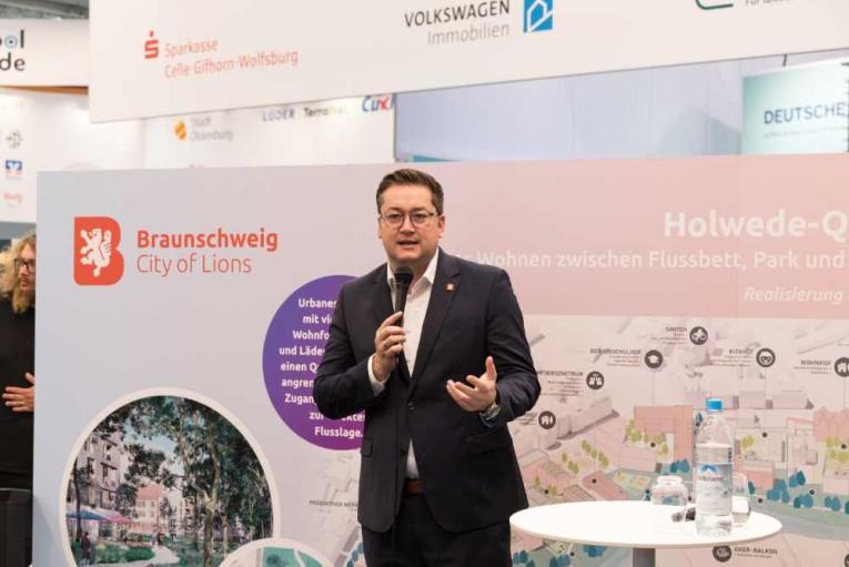 Mann im Anzug spricht mit Mikrofon vor einer Präsentationswand zur Stadtentwicklung in Braunschweig.