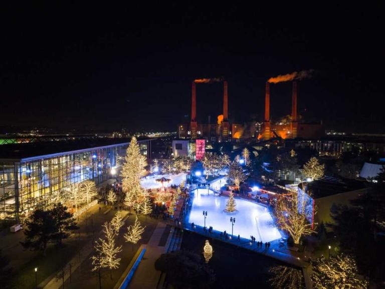 Nächtlicher Blick auf eine beleuchtete Eisbahn mit geschmückten Bäumen und Industrieanlagen im Hintergrund