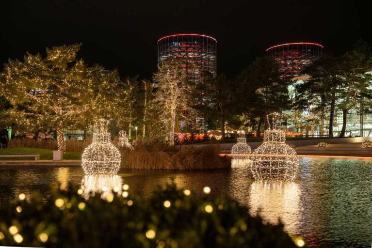 Nachtaufnahme eines beleuchteten Parks mit Lichtskulpturen im Wasser und Gebäuden im Hintergrund
